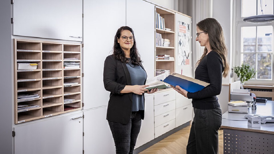 Zwei Frauen mit je einer Akte in der Hand stehen in einem Büro vor einem Schrank.