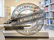 Ein Stempalaufdruck "Zusammen gegen  Rassismus - 100 % Menschenwürde" vor einem verschwommenen Blick in den Lesesaal "Anne-Frank-Shoah-Bibliothek" der Deutschen Nationalbibliothek in Leipzig